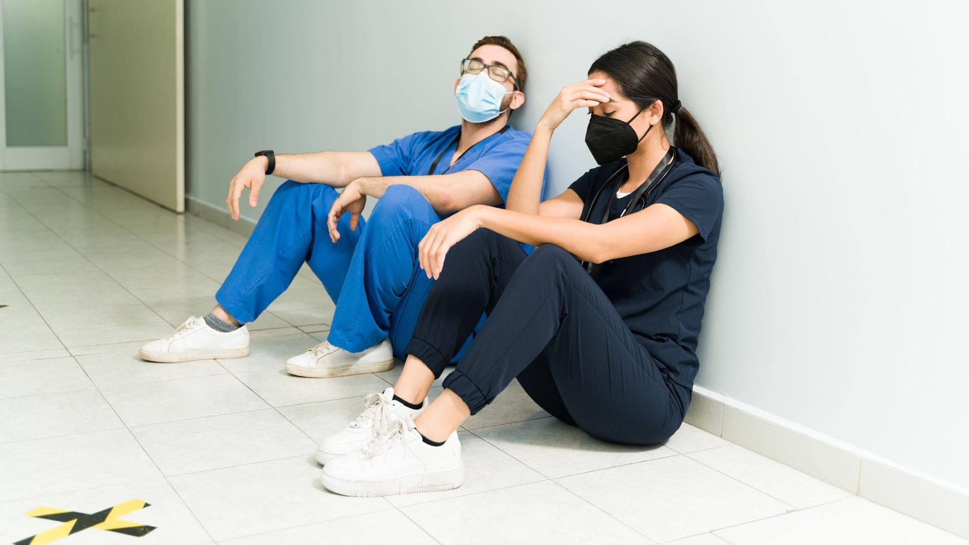 Two stressed out medical professionals, one a man and one a female, sit on the floor of a hospital.