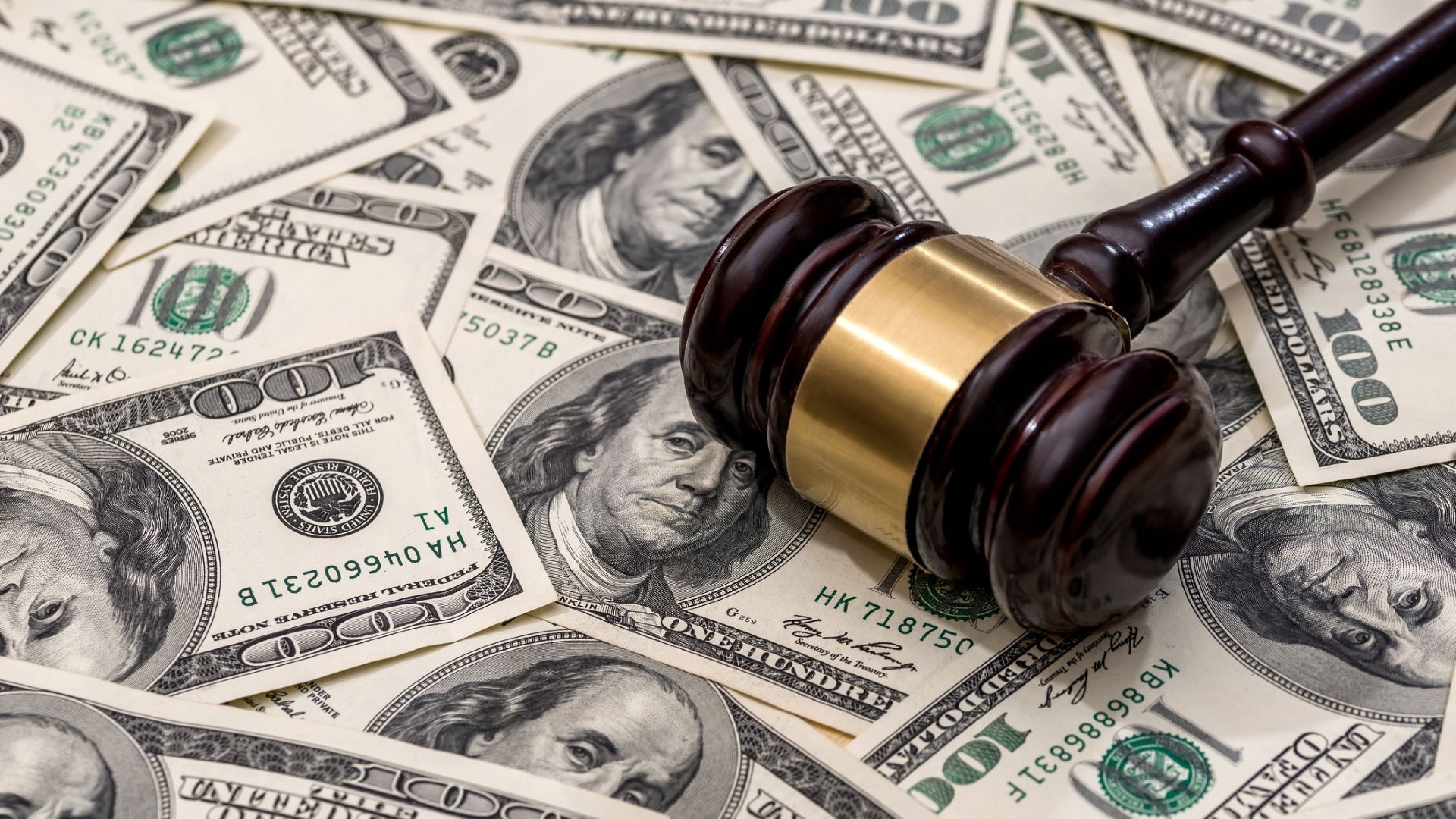 A spread out pile of hundred dollar bills with a judge's gavel on top of the pile.