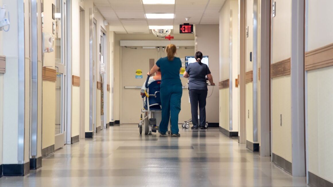 Two nurses walk down a hallway. One is pushing a patient that is sitting in a wheelchair while the other pushes a computer monitor.