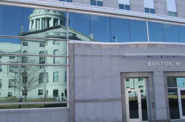 The State House building is reflected through the glass of the Cross office building