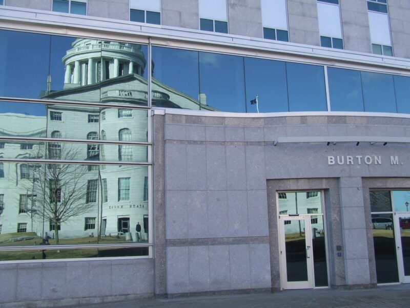 The State House building is reflected through the glass of the Cross office building