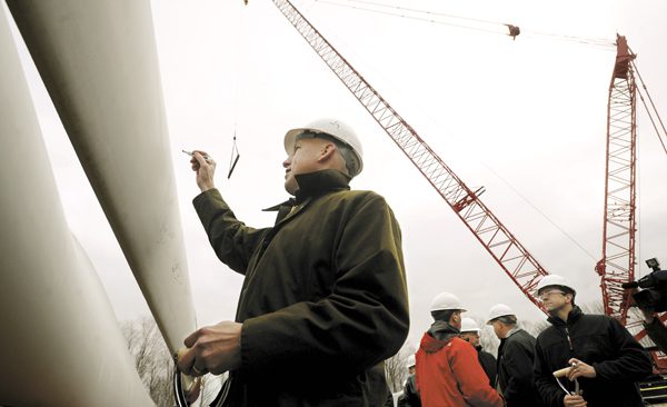 Paul Gaynor wearing a hard hat signs his signature to a blade of a windmill at a construction project