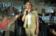 Cynthia Dill speaks into a microphone at a campaign event with a group of supporting, many holding blue signs with DILL on them standing behind her