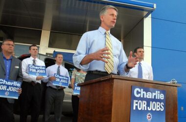 Charlie Summers speaks to a crowd of supporters during a campaign event while standing at a podium and talking into a microphone. A small group of supporters holding small blue signs stand behind him