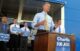 Charlie Summers speaks to a crowd of supporters during a campaign event while standing at a podium and talking into a microphone. A small group of supporters holding small blue signs stand behind him