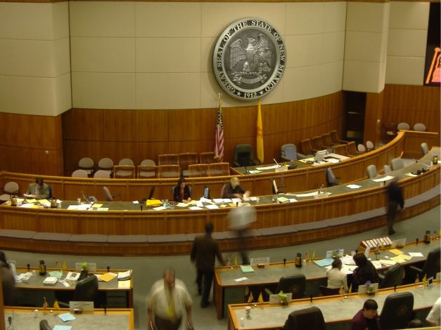 Overhead view of the inside of the New Mexico state legislature room filled with state lawmakers sitting at their desks and walking around.
