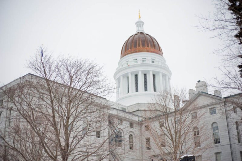 Exterior of the Maine State House during the winter with trees missing all their leaves.