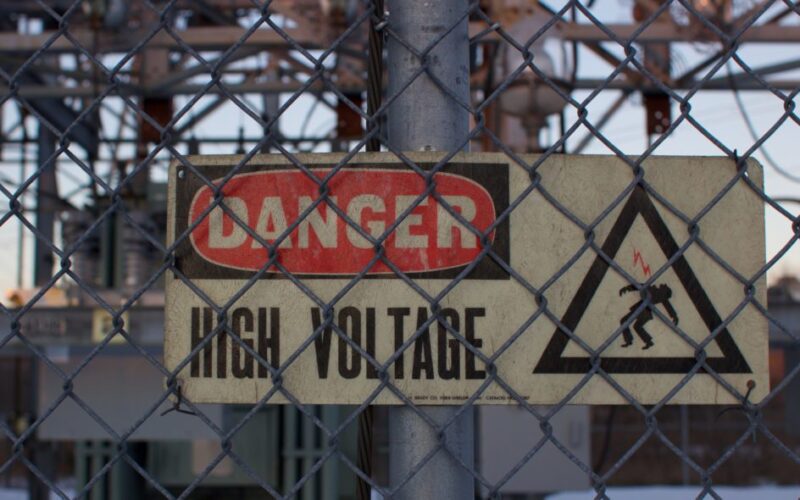 A sign reading DANGER HIGH VOLTAGE is affixed to a pole behind a chain link fence.