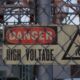 A sign reading DANGER HIGH VOLTAGE is affixed to a pole behind a chain link fence