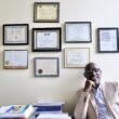 Claude Rwaganje laughs while sitting in a chair inside his office with a wall full of framed certificates behind him.