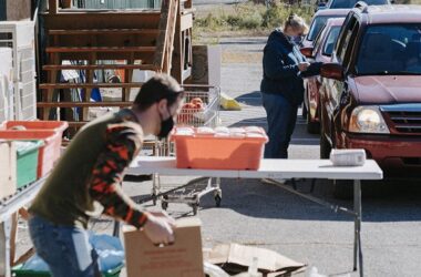 A man picks up a box at an outdoor food pantry and is beginning to carry the box to a car.