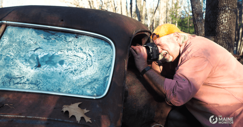 C. E. Morse leans ever so slightly into a junked car a junk yard with his camera to photograph the interior of the car.