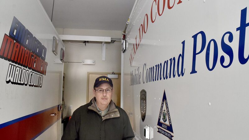 Emergency Management Director Dale Rowley stands within the agency's command center. To his left is a truck that reads "emergency management Waldo County" and to his right is a vehicle that reads "Waldo County Mobile Command Post."