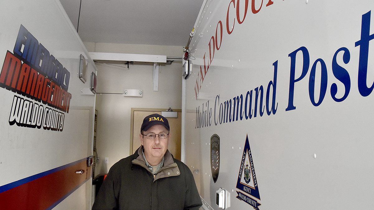 Emergency Management Director Dale Rowley stands within the agency's command center. To his left is a truck that reads "emergency management Waldo County" and to his right is a vehicle that reads "Waldo County Mobile Command Post."