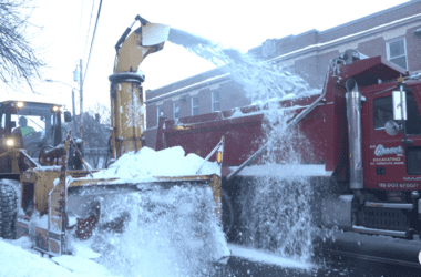 A snowplow deposits snow into the back of a dump truck.