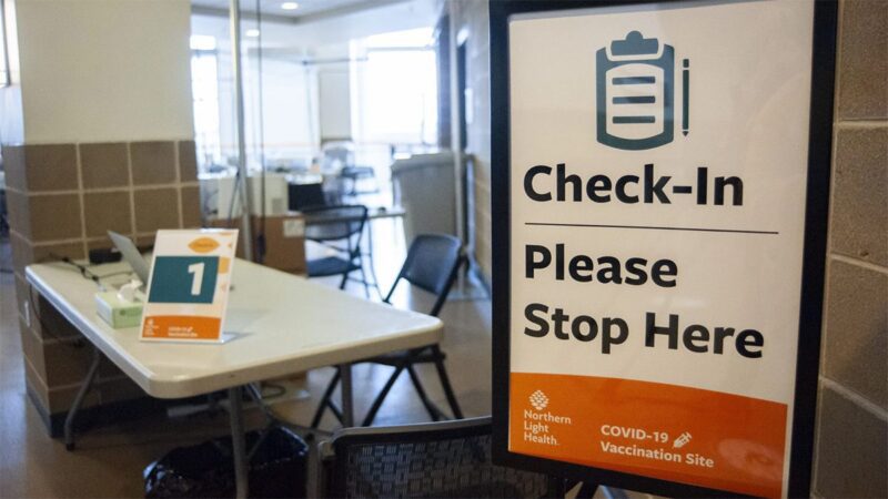 The entrance to a COVID-19 vaccination site in Bangor. Shown is the check-in table and a sign that reads "check-in please stop here. Northern Light Health COVID-19 vaccination site."