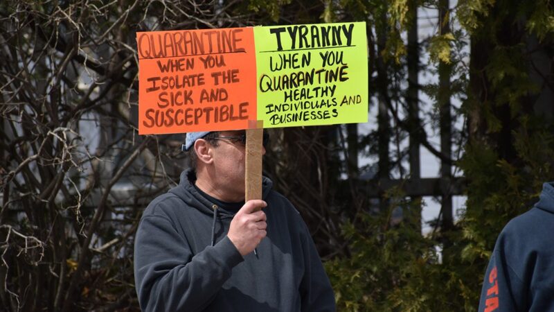 A man holds a sign during a protest outside the Maine State House. The reads: "Quarantine: When you isolate the sick and susceptible. Tyranny: When you quarantine health individuals and businesses."