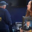 Bonnie Stewart looks off into the distance while holding a photo of her son. Her husband is in the background looking at a group of framed photos with his back towards the camera as he collects his composure.