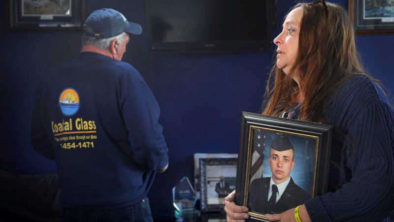 Bonnie Stewart looks off into the distance while holding a photo of her son. Her husband is in the background looking at a group of framed photos with his back towards the camera as he collects his composure.