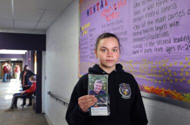 A young Washington County student holds a memoriam photo of classmate William McIver, who died by suicide, while standing in the hallway in front of a mental health resource display.