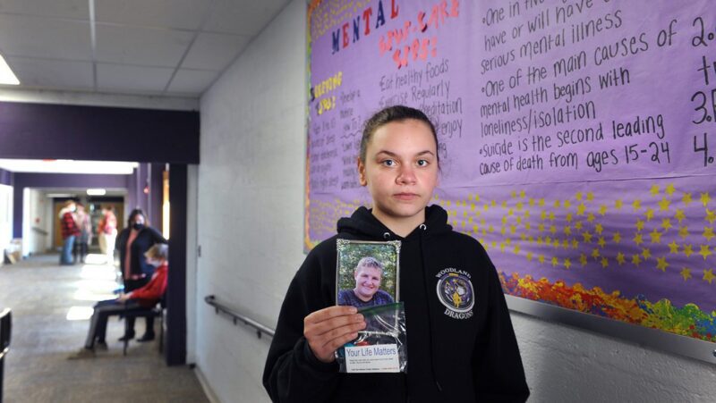A young Washington County student holds a memoriam photo of classmate William McIver, who died by suicide, while standing in the hallway in front of a mental health resource display.