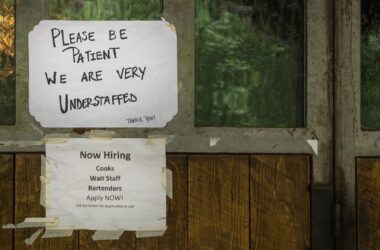 A pair of help wanted signs taped to the exterior of a building. One reads "Please be patient we are very understaffed, thank you" and the other reads "Now hiring cooks, wait staff, bartenders. Apply now! Ask bartender for application or call."
