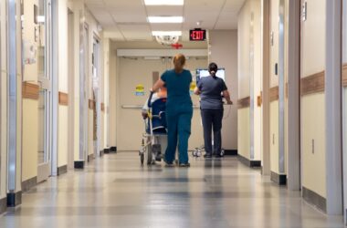 Two nurses walk down a hallway. One is pushing a patient that is sitting in a wheelchair while the other pushes a computer monitor.