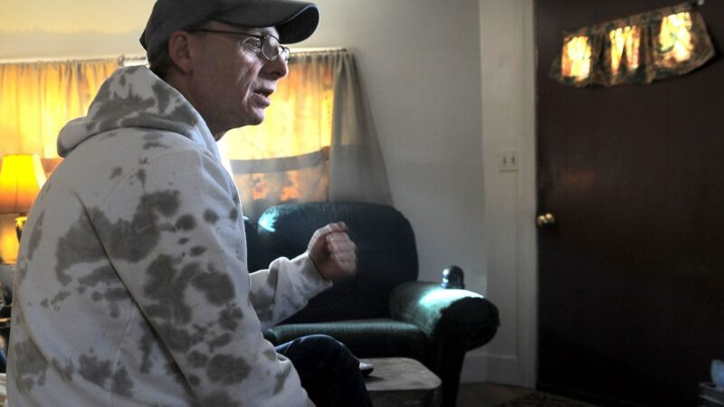 Steven Clarke sits in a motel room while he speaks with an individual not shown in the photo.
