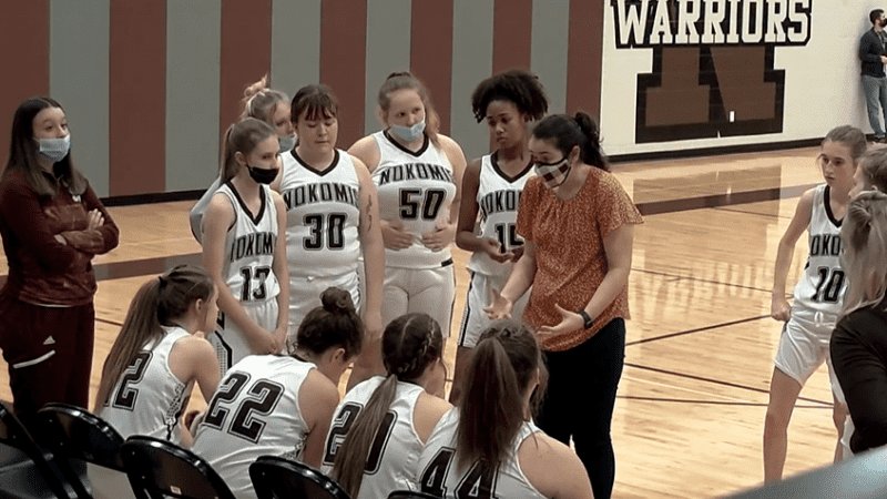A female basketball coach speaks to her basketball players in an open circle near the team bench