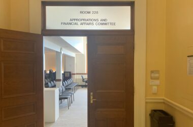 One of two doors leading into Room 228 in the state house, belonging to the Appropriations and Financial Affairs Committee, is open while the other door remains closed. Rows of empty chairs inside the room are visible