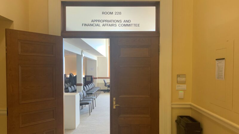 One of two doors leading into Room 228 in the state house, belonging to the Appropriations and Financial Affairs Committee, is open while the other door remains closed. Rows of empty chairs inside the room are visible