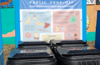 Four garbage cans sit in front of a banner that reads "public drop-off dump scraps from your own container" and shows what is accepted materials and what is forbidden materials. The items listed under each category are not legible in the photo.