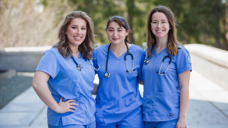 A group of three smiling women, each wearing nursing scrubs with a stethoscope around each of their necks