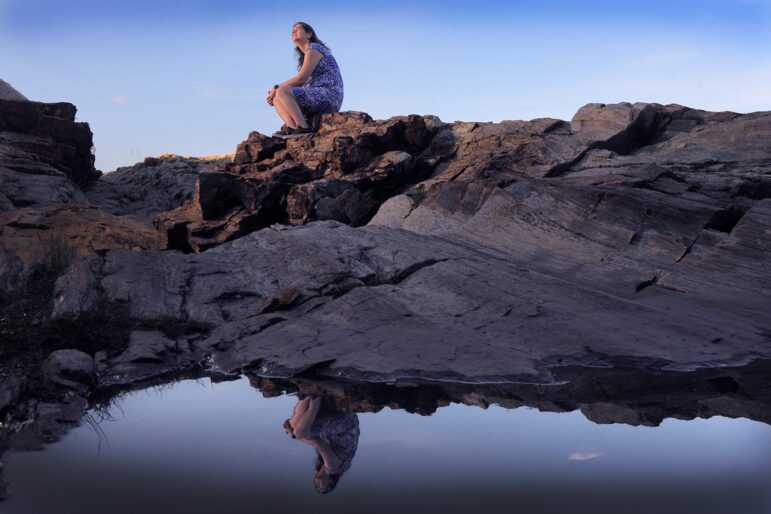 Deb McDonald sits on a large rock near water as she looks off into the distance