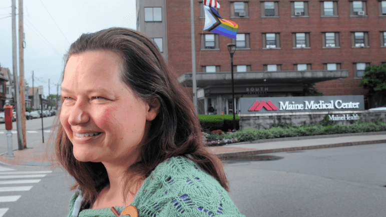 A nurse smiles as she stands across the street from the medical facility she works at with an LGTBQ flag flying on a flagpole