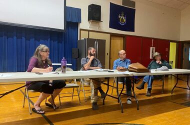 Members of the Columbia Falls planning and select boards sit at a table within the gymnasium and listen to a speaker that is not shown