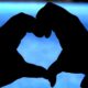 A silhouette of two hands forming the shape of a heart in front of a body of water