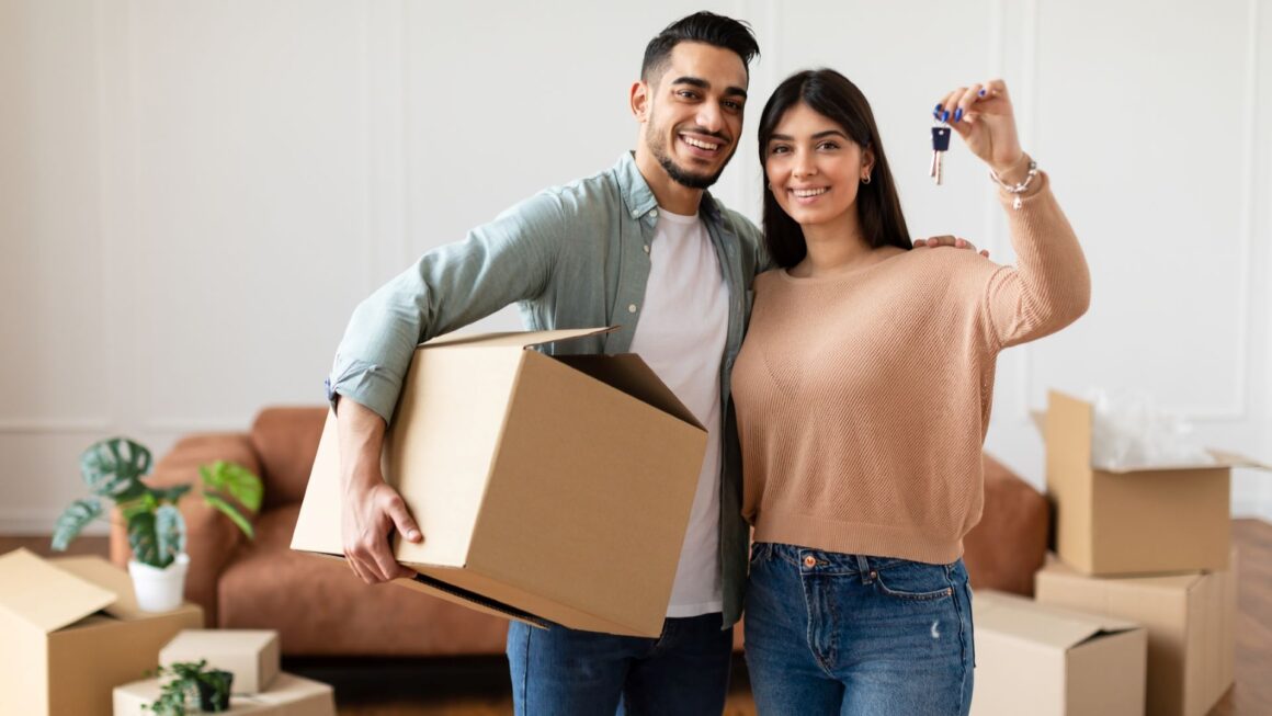 A man and a woman smile as they are in the process of moving homes. The man is holding a moving box tucked underneath his right arm with his left arm around the woman who is holding up the key to their new house in her left hand