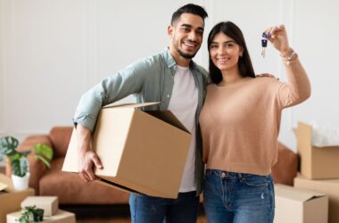 A man and a woman smile as they are in the process of moving homes. The man is holding a moving box tucked underneath his right arm with his left arm around the woman who is holding up the key to their new house in her left hand