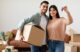 A man and a woman smile as they are in the process of moving homes. The man is holding a moving box tucked underneath his right arm with his left arm around the woman who is holding up the key to their new house in her left hand