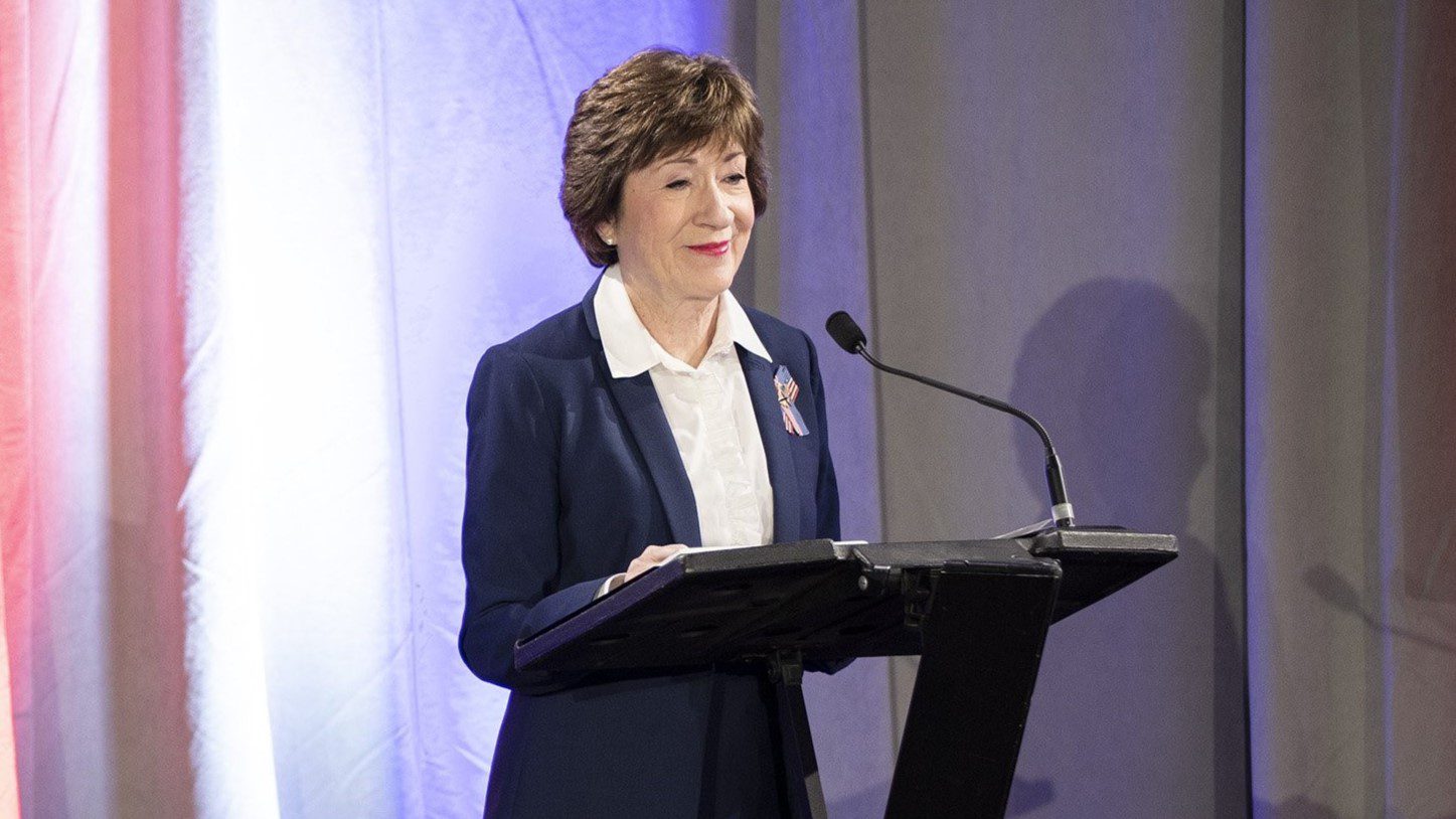 Susan Collins stands at a podium and prepares to speak into a microphone at an election debate