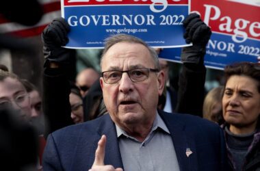 Paul LePage speaks during a campaign event with a crowd of people behind him, including some holding a red and blue sign that says LEPAGE GOVERNOR 2022