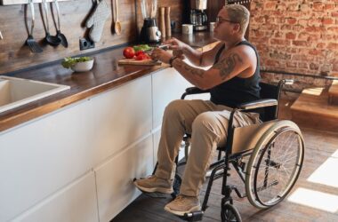 A woman sitting in a wheelchair cuts a tomato that is on a cutting board on her counter.