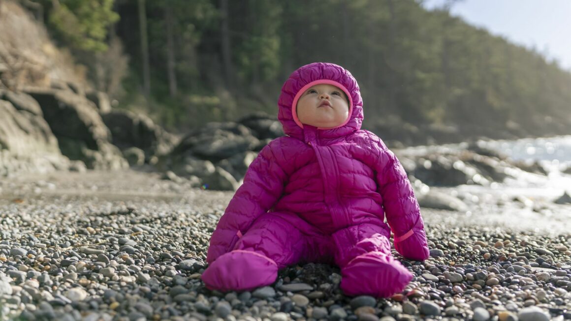 A baby in a puffy purple outdoor jacket sits on the rocks at a beach.
