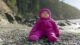 A baby in a puffy purple outdoor jacket sits on the rocks at a beach.