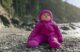 A baby in a puffy purple outdoor jacket sits on the rocks at a beach.