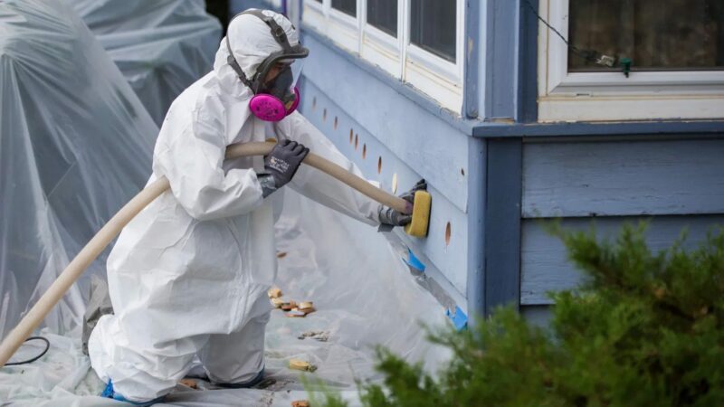 A man wearing a white protection suit injects insulation into the exterior wall of a home.