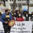 A man beats a drum in front of a medium-sized group of Indigenous people in support of a drinking water bill for Passamaquoddy Tribal members.