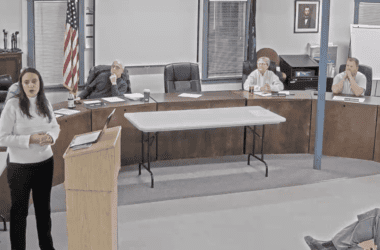 Shastri Ramnath, president of Exiro Mineral Corp, stands at a podium while speaking to a crowd of town residents. Members of the select board are seated behind her.