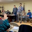 A speaker holding a microphone stands at the front of the town meeting and addresses the seated residents
